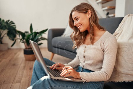 Téléchargez les photos : Jeune femme travaillant sur un ordinateur portable tout en étant assis sur le sol près du canapé dans le salon à la maison. Travail à distance et freelance. - en image libre de droit