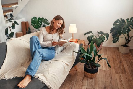Photo for Young woman reading a book on the sofa in a cozy living room at home relaxing and unwinding on her day off. - Royalty Free Image