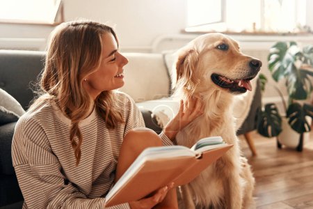 Foto de Mujer joven feliz con un perro en el sofá en una acogedora sala de estar en casa relajante y descansando en un día libre, abrazando y jugando con su mascota y leyendo un libro. - Imagen libre de derechos