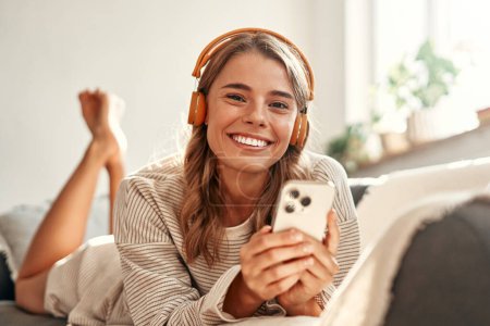 Téléchargez les photos : Joyeux jeune femme dans les écouteurs avec téléphone sur canapé dans le salon confortable à la maison relaxant et se détendre le week-end. - en image libre de droit