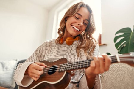 Téléchargez les photos : Joyeux jeune femme jouant de la guitare tout en étant assis sur le canapé dans le salon confortable à la maison relaxant et se détendre le week-end. - en image libre de droit