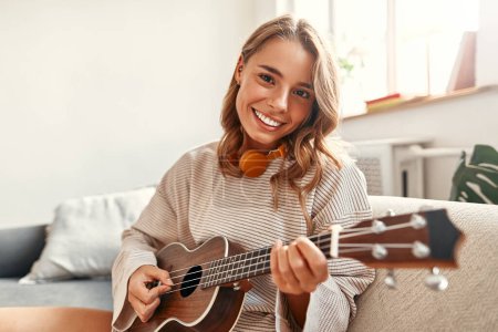 Téléchargez les photos : Joyeux jeune femme jouant de la guitare tout en étant assis sur le canapé dans le salon confortable à la maison relaxant et se détendre le week-end. - en image libre de droit