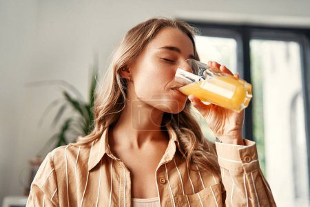 Foto de Una joven mujer feliz parada en la cocina bebe jugo de naranja de un vaso. Mujer desayunando en una acogedora cocina. - Imagen libre de derechos