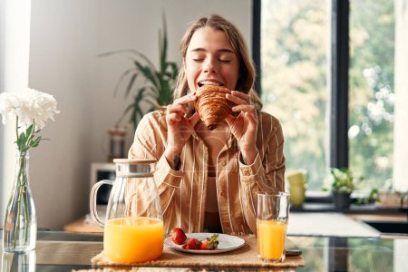 Foto de Joven mujer feliz sentada en la mesa de la cocina, comiendo croissants con fresas y bebiendo jugo. Mujer desayunando en una acogedora cocina. - Imagen libre de derechos