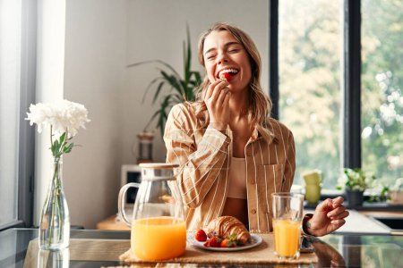 Photo for Young happy woman sitting at the table in the kitchen, eating croissants with strawberries and drinking juice. Woman having breakfast in a cozy kitchen. - Royalty Free Image