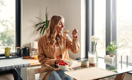 Foto de Joven mujer feliz sentada a la mesa en la cocina, comiendo cereales con leche, fresas y tomando café. Una mujer está desayunando en una acogedora cocina. - Imagen libre de derechos