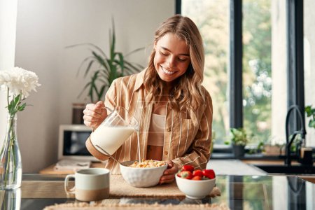 Photo for A woman preparing herself breakfast, pouring milk into a plate with cereal, strawberries and a cup of coffee standing nearby. A woman is having breakfast in a cozy kitchen. - Royalty Free Image