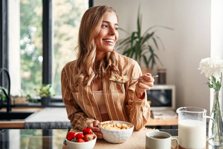 Foto de Joven mujer feliz sentada a la mesa en la cocina, comiendo cereales con leche, fresas y tomando café. Una mujer está desayunando en una acogedora cocina. - Imagen libre de derechos