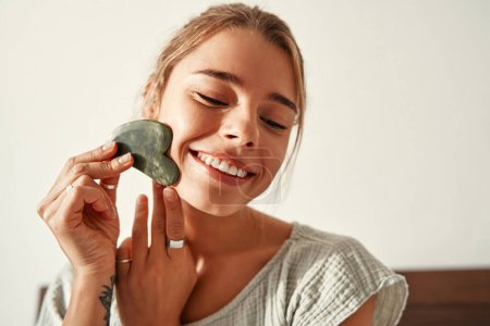 Foto de Una joven mujer feliz despertando por la mañana se sienta en la cama, haciendo un masaje facial con un raspador de jade gua sha. Cuidado de la piel para la cara y el cuerpo, rutina matutina, tratamientos de spa. - Imagen libre de derechos