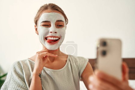 Téléchargez les photos : Une femme a appliqué un masque facial en argile le matin et a pris un selfie sur un smartphone dans la chambre à coucher à la maison. Soins de la peau et soins spa. - en image libre de droit