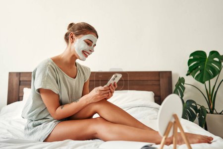 Téléchargez les photos : Une femme a appliqué un masque facial en argile le matin alors qu'elle était assise avec un smartphone sur le lit dans la chambre à coucher à la maison. Soins de la peau et soins spa. - en image libre de droit