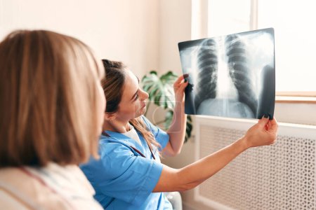 Téléchargez les photos : Une femme médecin de famille à la maison dans un uniforme médical avec un stéthoscope examinant une radiographie des poumons d'une femme âgée alors qu'elle était assise sur un canapé dans le salon. Soins de santé et médecine. - en image libre de droit