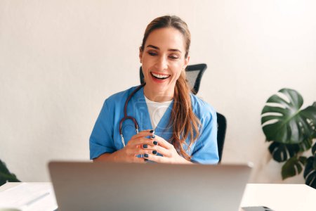 Photo for Woman doctor at home in medical uniform with stethoscope sitting at table with laptop talking on video call in living room. Health care and medicine, online consultation. - Royalty Free Image