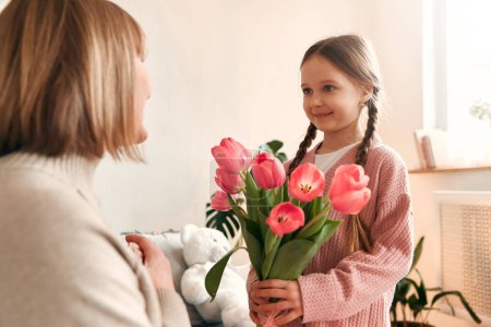 Photo for Cute child granddaughter congratulating her grandmother by giving her flowers and a gift while sitting on the sofa in the living room. Concept of Women's Day and Mother's Day. - Royalty Free Image