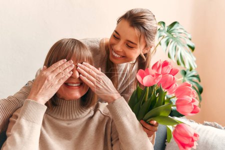 Photo for Young woman congratulating her grandmother or mother by giving her flowers and a gift while sitting on the sofa in the living room. Mother's day and women's day concept. - Royalty Free Image