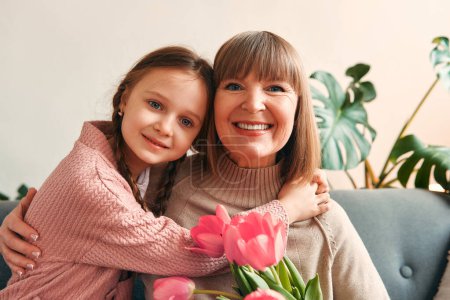 Foto de Linda nieta felicitando a su abuela dándole flores mientras está sentada en el sofá de la sala de estar. Día de la madre y concepto del día de la mujer. - Imagen libre de derechos