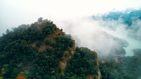 Téléchargez les photos : Un paysage naturel envoûtant avec une montagne entourée de brouillard et d'arbres, un lac serein en arrière-plan, sous un ciel nuageux - en image libre de droit