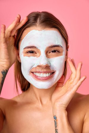 Photo for Beautiful happy woman using cleansing clay face mask for blackheads and enlarged pores isolated on pink background. Skin care, cosmetology, beauty concept. - Royalty Free Image