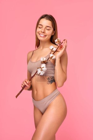 Schöne Frau in Dessous mit Baumwollzweig auf rosa Hintergrund. Körperpflege, Kosmetologie, Schönheitskonzept.