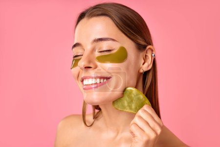 Beautiful woman with moisturizing patches under her eyes doing massage with gua sha scraper isolated on pink background. Facial skin care, cosmetology, beauty concept.