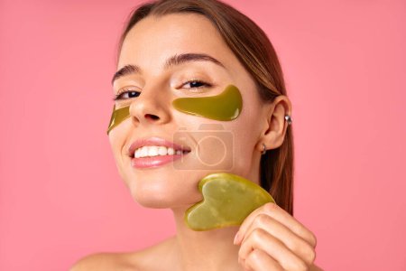 Photo for Beautiful woman with moisturizing patches under her eyes doing massage with gua sha scraper isolated on pink background. Facial skin care, cosmetology, beauty concept. - Royalty Free Image