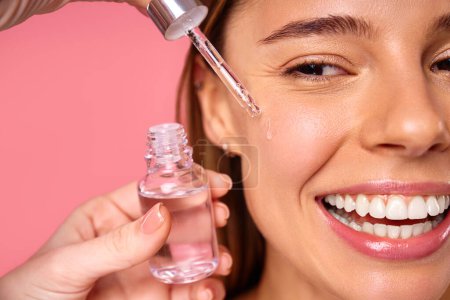 Beautiful woman face, natural clean and fresh glowing skin, applying anti-aging collagen serum. Hands hold serum with active herbal ingredients vitamin C in a dropper, on a pink background.