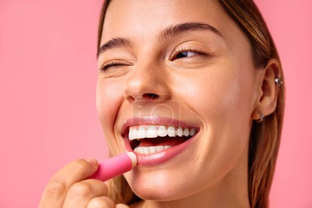 Beautiful well-groomed woman with clean glowing healthy skin using moisturizing lip balm isolated on pink background. Facial skin care, cosmetology, beauty concept.