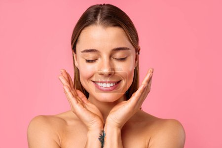 A cheerful young woman with tattoos on her arms is smiling and gently touching her face while posing against a pink background. The image conveys a theme of radiantly happy skin care