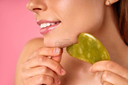 Eine Frau gönnt sich Selbstpflege und Beauty-Routine mit einer Jade-Gesichtswalze am Kinn, die ihre Haut verwöhnt. Gelassenes Ambiente mit rosa Hintergrund stimmt auf eine entspannende Hautpflege-Sitzung ein