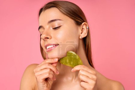 A woman indulges in selfcare and beauty routine with a jade facial roller on her chin, pampering her skin. Serene ambiance with a pink background sets the mood for a relaxing skincare session