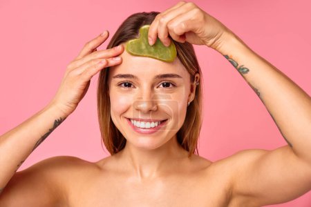 A woman indulges in selfcare and beauty routine with a jade facial roller on her chin, pampering her skin. Serene ambiance with a pink background sets the mood for a relaxing skincare session