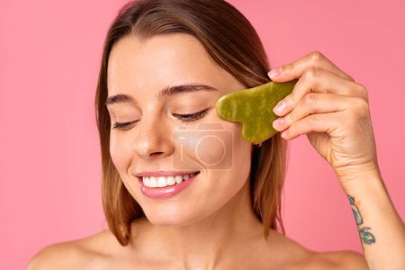 Eine Frau gönnt sich Selbstpflege und Beauty-Routine mit einer Jade-Gesichtswalze am Kinn, die ihre Haut verwöhnt. Gelassenes Ambiente mit rosa Hintergrund stimmt auf eine entspannende Hautpflege-Sitzung ein