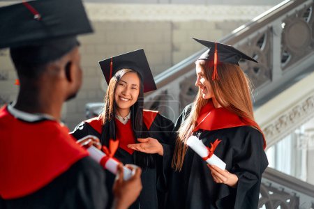 Vielfältige Gruppe glücklicher Studenten in Abschlusskleidern und -mützen feiern akademische Leistungen mit einem Lächeln bei freudiger Zeremonie, Bindung und Gratulation bei Meilenstein-Erfolg