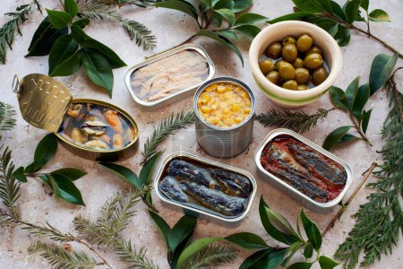 Various tins of seafood and a bowl of olives, surrounded by fresh green leaves.