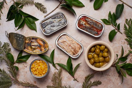 Various tins of seafood and a bowl of olives, surrounded by fresh green leaves.