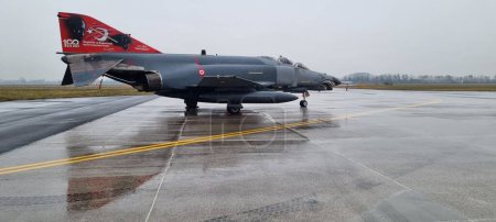 Photo for Turkish F-4 Phantom fighter jet parked on a rainy day, its formidable presence underscored by the relentless downpour, capturing a moment of quiet strength. - Royalty Free Image