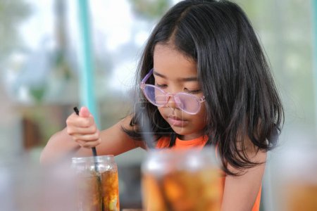 Téléchargez les photos : Expression faciale de petite fille assise seule à boire du thé au citron glacé - en image libre de droit