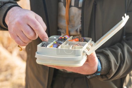 Les mains du pêcheur tiennent une boîte en plastique avec une variété de leurres et de crochets de pêche.