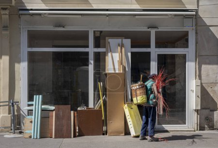 Téléchargez les photos : Paris, France - 05 11 2024 : Vue d'une vieille glaneuse portant une cagoule sur le dos devant des objets encombrants dans un magasin fermé - en image libre de droit