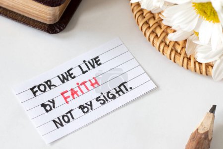 Photo for We live by faith, not by sight, handwritten Christian quote on paper with pencil, flowers, and closed holy bible book. Biblical concept of trust in God Jesus Christ, 2 Corinthians 5:7 text. - Royalty Free Image