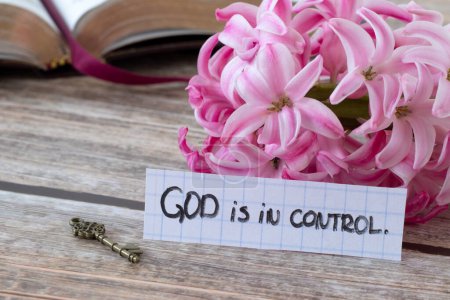 God is in control, handwritten quote with flower, ancient key, and open holy bible book on wood. Close-up. Christian biblical concept of purpose, comfort, guidance, protection, faith in Jesus Christ.