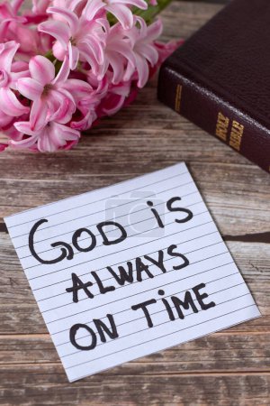 God is always on time, inspirational handwritten quote with holy bible and flowers on wood. Christian biblical concept of patience, waiting on Jesus Christ, answered prayer, and faith.