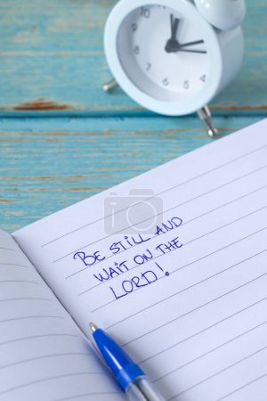 Be still and wait on the LORD, handwritten quote in notebook with alarm clock on wooden table. Close-up. Biblical concept of Christian time, patience, peace, trust, and faith in God Jesus Christ.