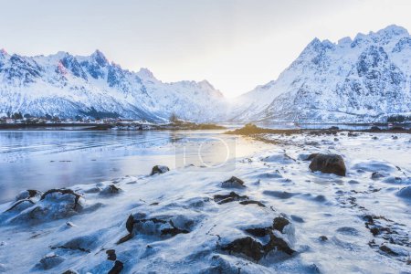 Téléchargez les photos : Belles montagnes enneigées avec de la glace au premier plan et un soleil doré au lever du soleil en hiver dans les îles Lofoten, Norvège. - en image libre de droit