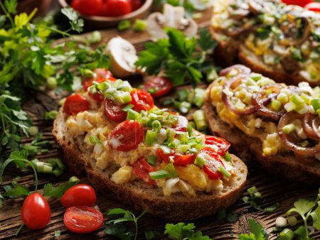 Photo for Close up on sandwich with scrambled eggs, cherry tomatoes and chopped green onion on a wooden board among the fresh herbs - Royalty Free Image
