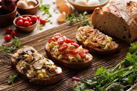 Foto de Sandwiches con huevos revueltos y verduras en una tabla de madera con aros de cebolla en una tabla de madera - Imagen libre de derechos