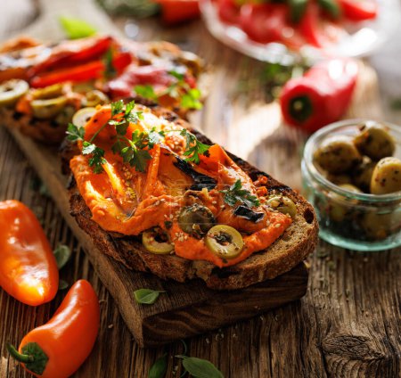 Téléchargez les photos : Sandwich au pain au levain traditionnel grillé au houmous de tomate, poivrons grillés et olives vertes saupoudrés de persil frais sur une planche en bois, vue rapprochée - en image libre de droit