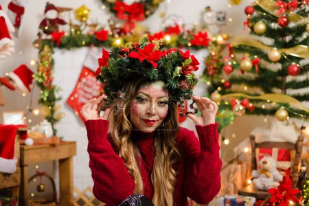 Téléchargez les photos : Belle femme avec maquillage de Noël portant une couronne de Noël et un pull rouge assis près de la cheminée dans le salon décoré pour Noël, tenant un coupe-biscuits. - en image libre de droit