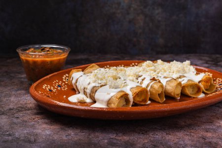 Photo for Fried potato tacos with cream and cheese in a Mexican mud dish. Tacos dorados, Mexican food. - Royalty Free Image