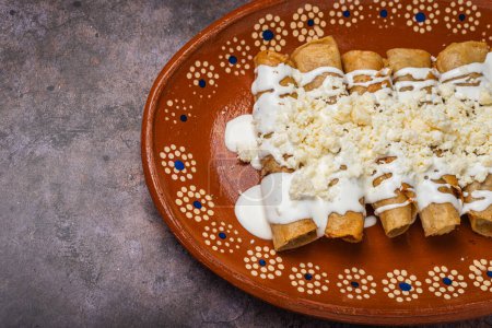 Photo for Fried potato tacos with cream and cheese in a Mexican mud dish. Tacos dorados, Mexican food. - Royalty Free Image